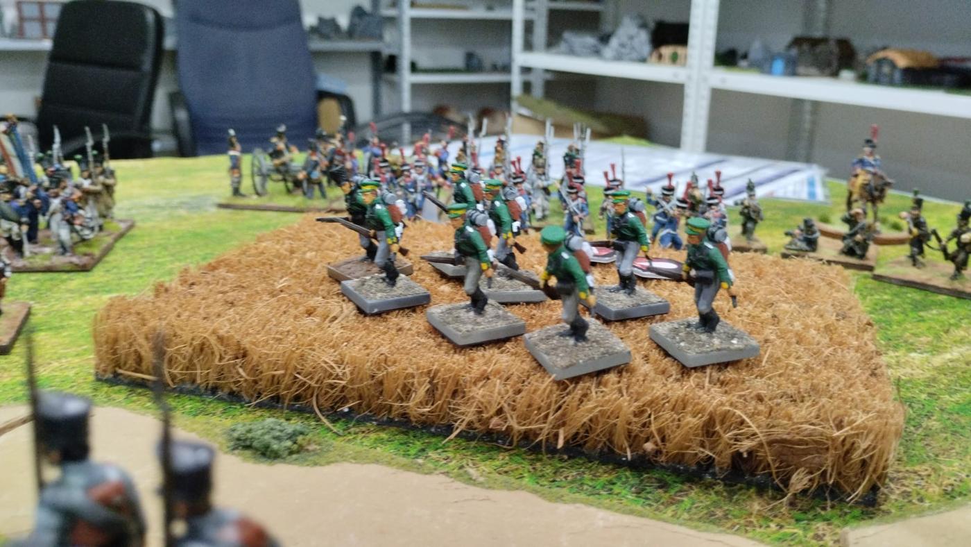 Cazadores prusianos de uniforme verde avanzando por un trigal, con infantería y artillería francesas al fondo