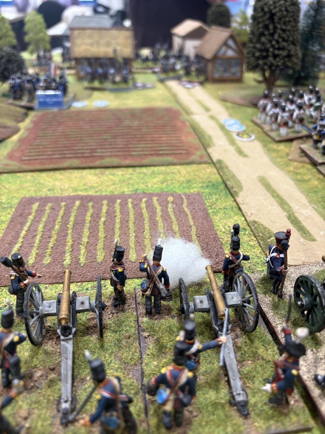 Primer plano de una batería francesa disparando, con humo de pólvora, frente a los campos y el pueblo